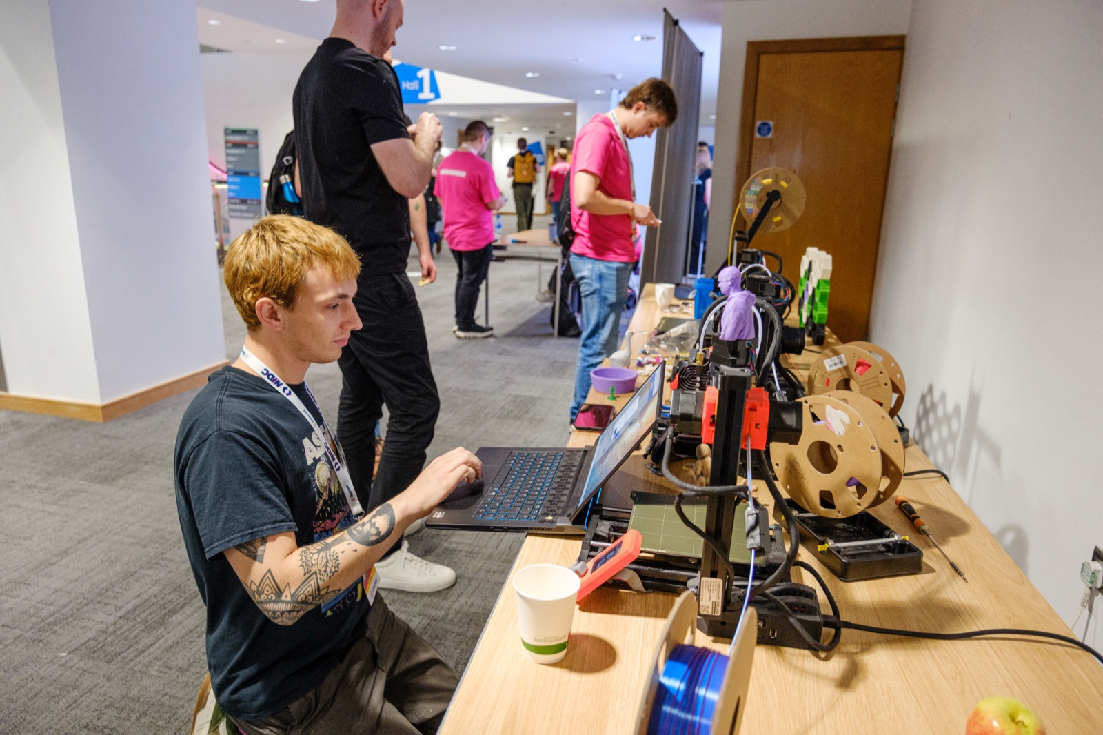An attendee works at a laptop beside a desktop 3D printer and reels of filament at an NIDC village stand